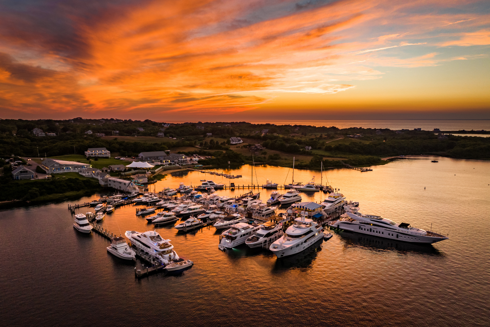 A marina filled with luxury yachts docked at sunset, calm waters reflecting orange skies, a peaceful coastal village in the distance, golden light.